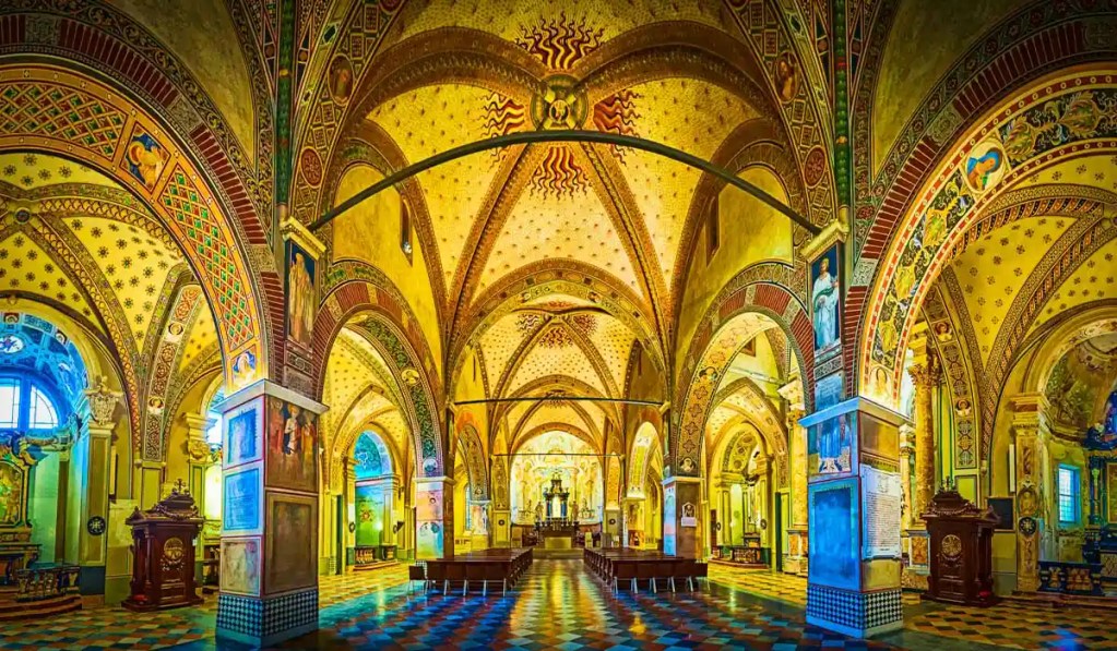 The ornate interior of the Cathedral of San Lorenzo in Lugano, Switzerland, showcasing intricate frescoes and vibrant patterns adorning the arched ceilings and walls. The central aisle leads to the main altar, surrounded by detailed artwork and illuminated by natural light streaming through the windows.