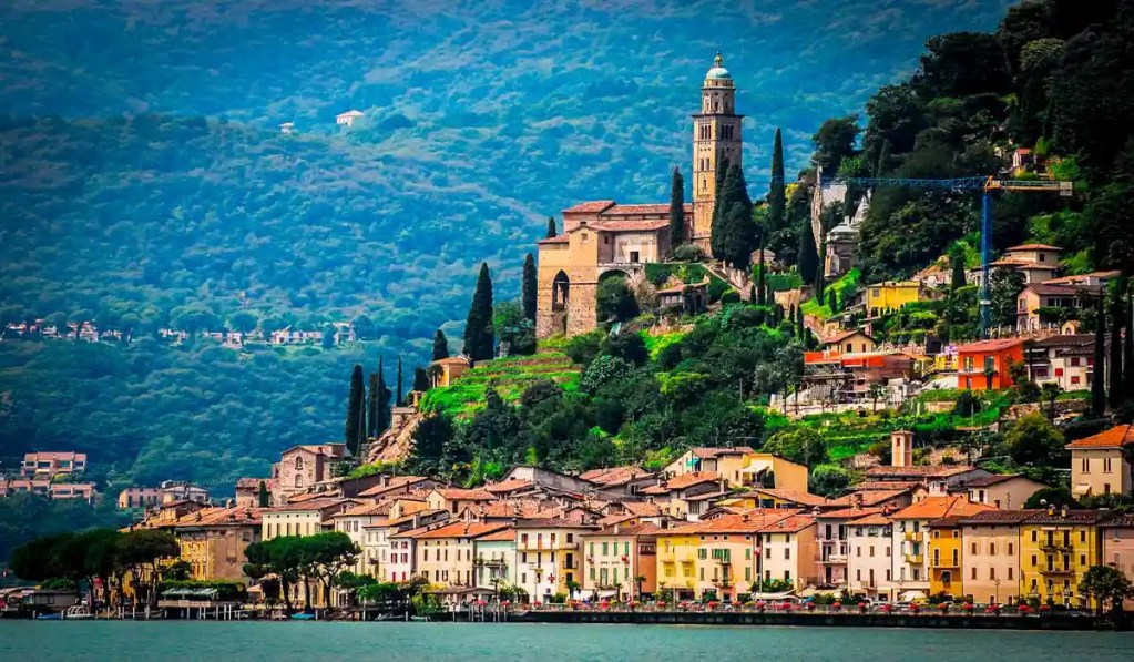 A picturesque view of Morcote, Switzerland, featuring colorful houses lining the waterfront with a hillside backdrop. The village is crowned by the historic Santa Maria del Sasso church, surrounded by tall cypress trees and lush greenery, with the serene waters of Lake Lugano in the foreground.