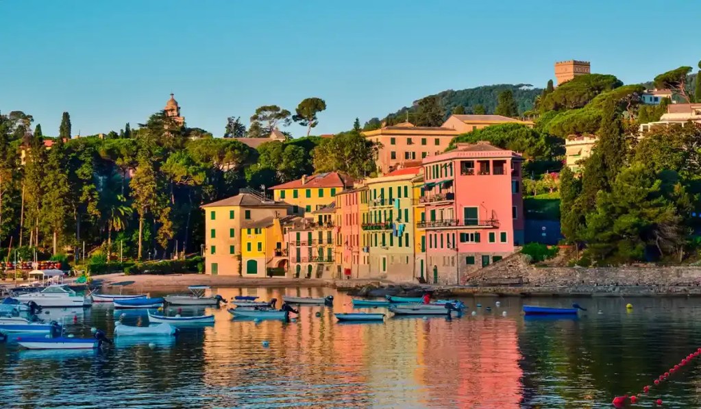 This image depicts the serene waterfront of San Michele di Pagana. Vibrantly colored buildings line the shore, their reflections shimmering on the calm water, while small boats float peacefully in the foreground. The backdrop features lush greenery and historic architecture, including a castle-like structure atop a hill. 