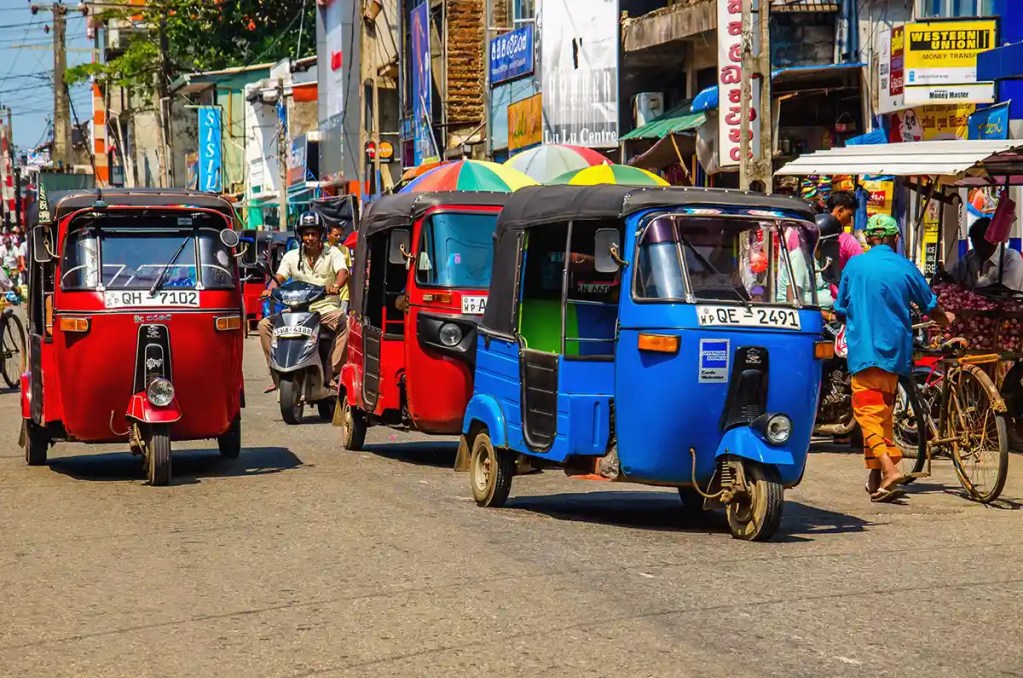 A bustling street scene with colorful tuk-tuks in red and blue navigating through traffic alongside a motorbike and pedestrians. The vibrant backdrop features busy market stalls, signboards, and a mix of urban activity, capturing the lively atmosphere of a Sri Lankan marketplace.