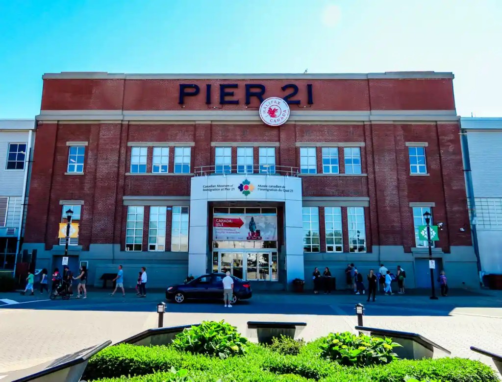 Front view of the Canadian Museum of Immigration at Pier 21 in Halifax, featuring a red brick facade with large windows, bold "PIER 21" signage across the top, and people walking and gathering outside the entrance under a sunny blue sky.