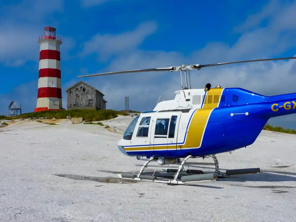 A blue, white, and yellow helicopter is parked on a rocky shoreline with a tall red-and-white striped lighthouse and a weathered wooden building in the background, under a bright blue sky with scattered clouds.