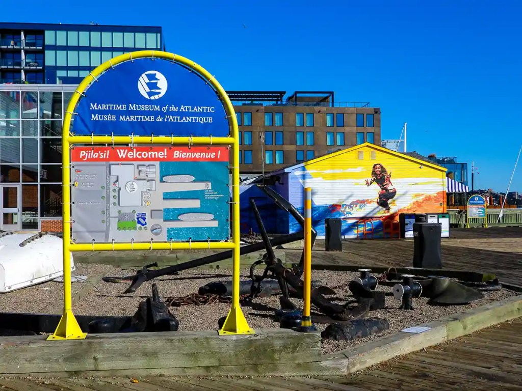 The entrance to the Maritime Museum of the Atlantic shows a large yellow and blue welcome sign with a site map, flanked by maritime artifacts like anchors and chains; in the background, a bright yellow building features a mural of a woman in a wetsuit jumping joyfully over stylized waves.