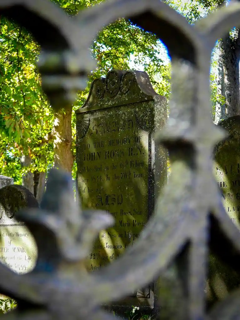 A close-up view through an ornate wrought iron fence shows aged, moss-covered gravestones in a historic cemetery, with dappled sunlight filtering through green leafy trees overhead.