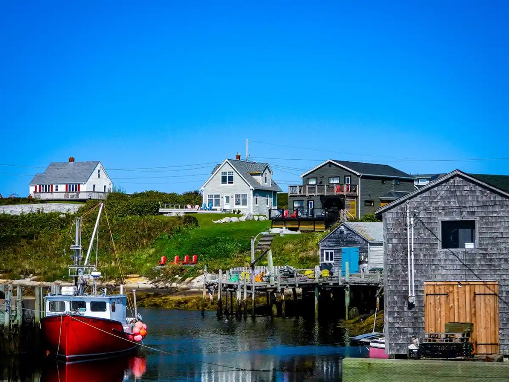A small harbor at Peggy’s Cove features a bright red fishing boat moored alongside weathered wooden docks, with colorful coastal houses and a clear blue sky in the background.
