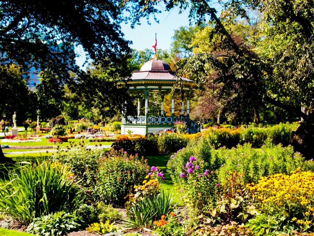 Colorful scene from the Halifax Public Gardens featuring a historic bandstand surrounded by vibrant flower beds, lush green lawns, and mature trees casting dappled shadows under a bright, sunny sky.