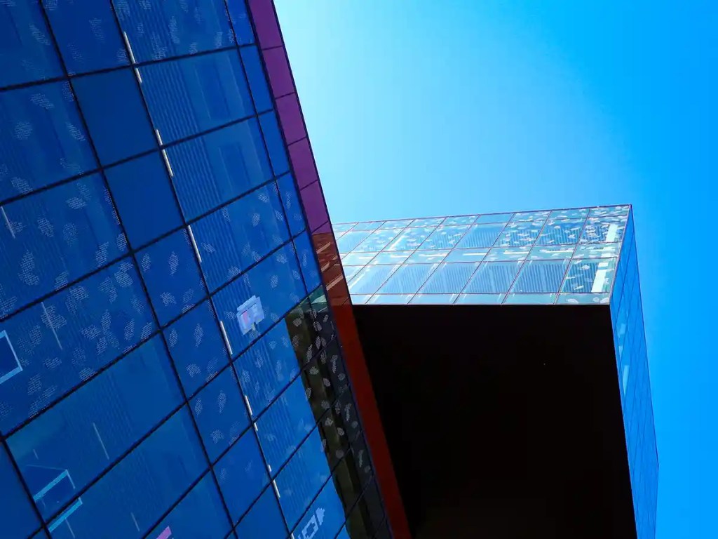 An angled view looking up at the modern glass facade of the Halifax Central Library, reflecting the clear blue sky and showing detailed leaf patterns on the windows.