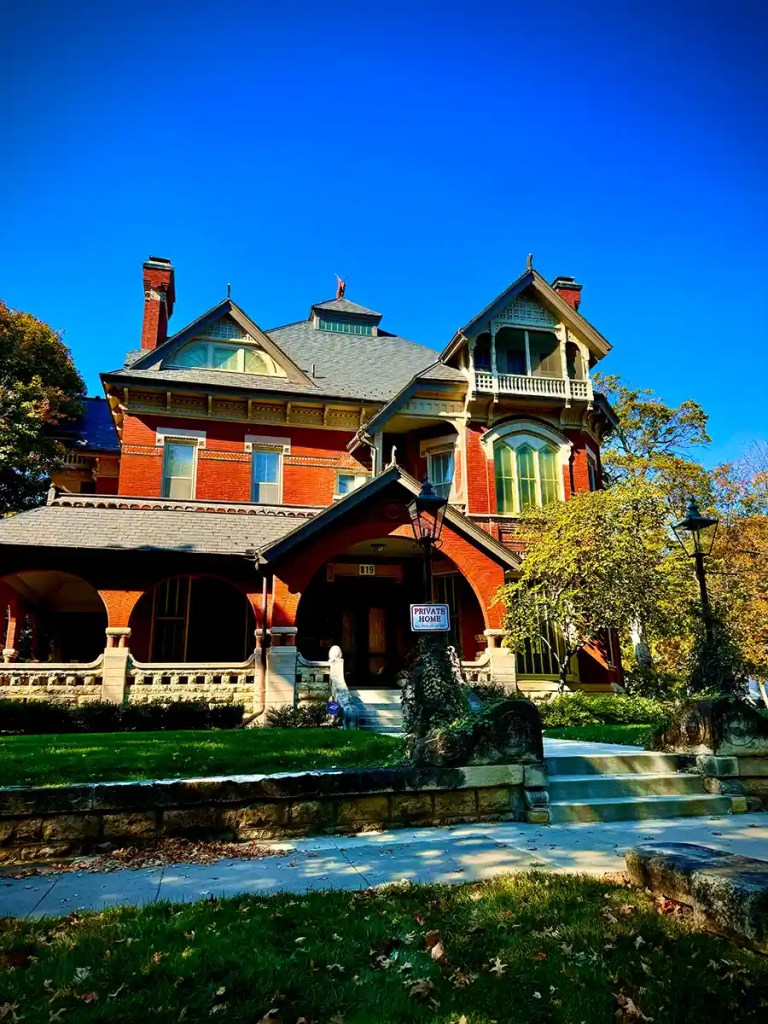 A grand, multi-story Victorian-style brick house with ornate architectural details, arched porchways, and a steeply pitched roof, set against a vibrant blue sky. A sign reading “Private Home” is posted at the front steps, which are flanked by stone columns and lush green landscaping.