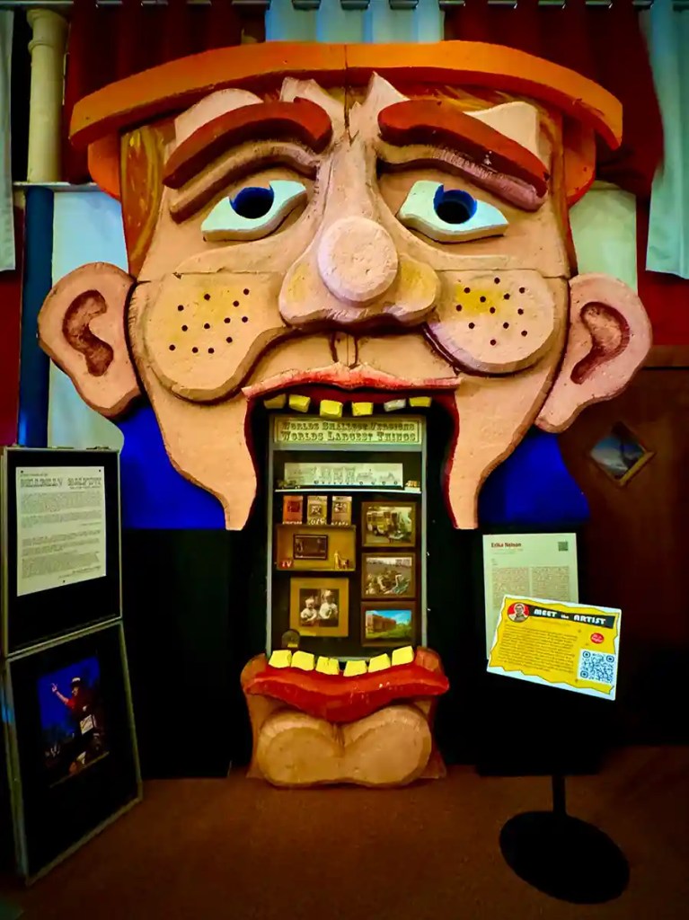 A large, cartoonish face sculpture with exaggerated features—freckles, red eyebrows, and a wide open mouth—frames a display case set inside the mouth, giving the illusion that the viewer is looking into the character’s throat. The display includes miniature dioramas and signage labeled “World’s Largest Things,” with informational plaques and artwork panels on either side.