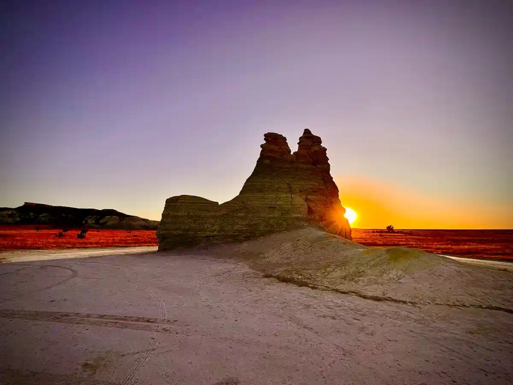 A jagged rock formation stands tall in a flat, open landscape as the sun sets directly behind it, casting a golden glow and long shadows across the ground. The sky fades from deep purple to orange, creating a dramatic contrast with the red and brown tones of the surrounding plains.