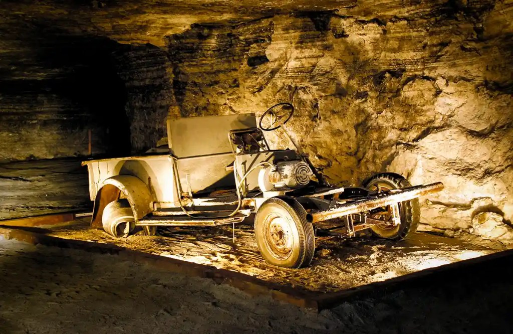 An old, rusted mining vehicle with missing parts and exposed mechanical components is displayed in a dimly lit underground cavern. The rough, stratified rock walls surrounding it highlight the vehicle’s historical use in subterranean environments.