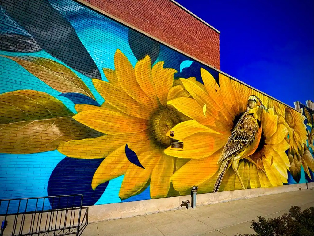 A vibrant mural painted on a brick wall features large, detailed sunflowers in full bloom with a realistic yellow and brown bird perched on one of the blossoms. The bright blue background enhances the bold contrast of the yellow petals, creating a lively and inviting streetscape scene.