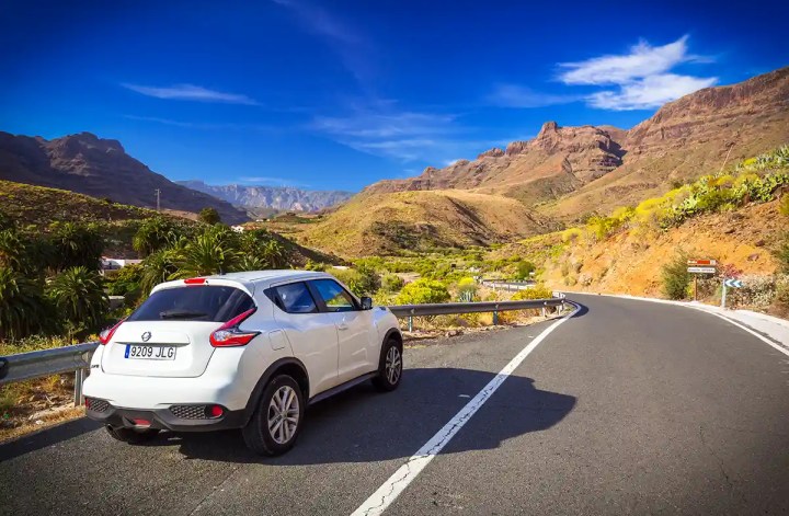 A white SUV parked on the shoulder of a winding mountain road under a vivid blue sky, surrounded by arid hills, cacti, and palm trees. The scenic landscape features rugged peaks and lush greenery, hinting at a warm, sunny climate ideal for a road trip to Andorra.