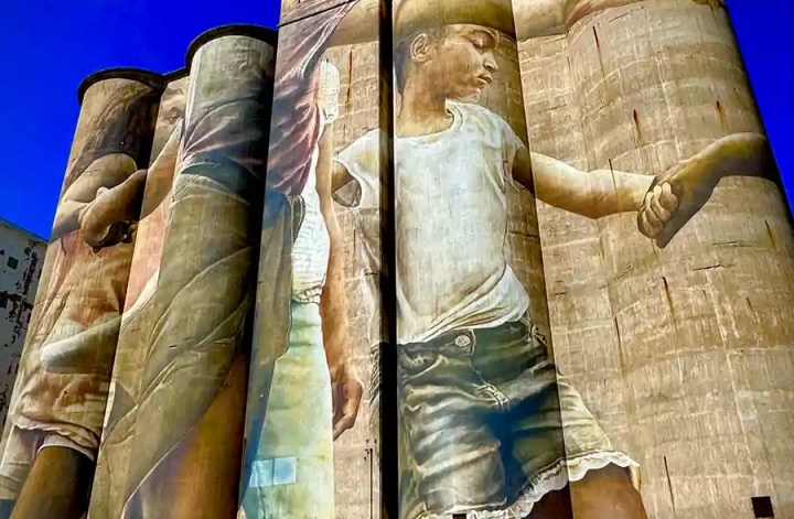 A towering mural painted across several grain silos depicts children holding hands, rendered in soft, lifelike tones. The artwork contrasts beautifully against the deep blue sky, transforming the industrial structure into a striking public art installation.