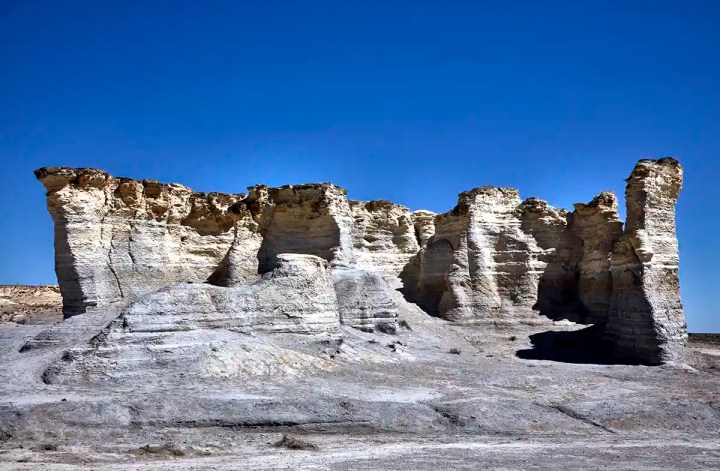 Wide view of a weathered limestone wall formation under a deep blue sky, featuring tall, eroded spires and striated layers. The light-colored rock contrasts with the barren, dusty ground, showcasing the dramatic natural landscape of the prairie badlands.