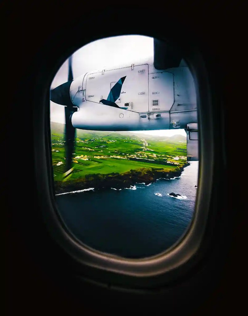 View through an airplane window showing a propeller engine in motion and a vibrant coastline below, with lush green fields and clusters of small houses near the rocky shore. The dark ocean water contrasts with the bright, patchwork farmland.