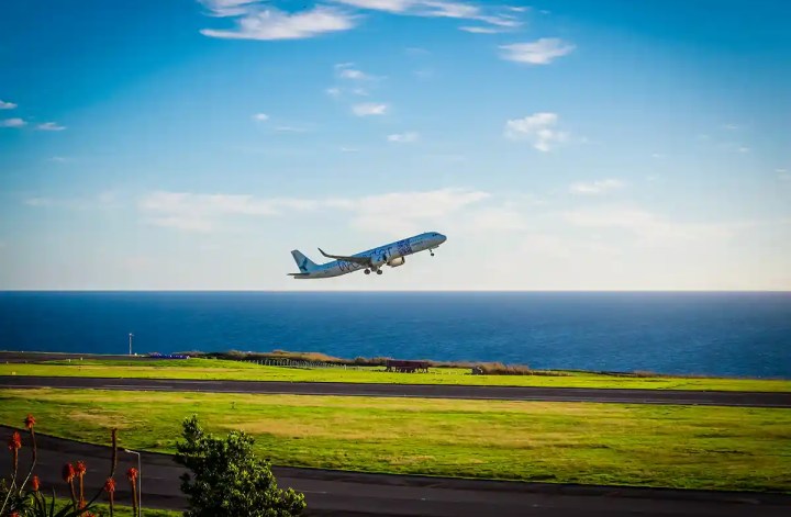 A commercial airplane takes off from a coastal runway, climbing into a bright blue sky with the ocean stretching out behind it. The green grass of the airfield contrasts sharply with the deep blue sea and sky.