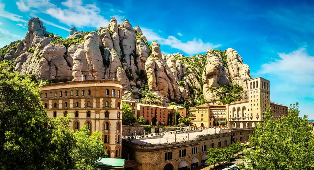 This image showcases the Montserrat Monastery nestled against dramatic, jagged limestone peaks in Catalonia, Spain. Historic stone buildings with arched windows and terraced roofs sit beneath the towering rock formations, surrounded by lush greenery and a bright blue sky. The scene highlights the stunning natural and architectural contrast of this famous pilgrimage site.