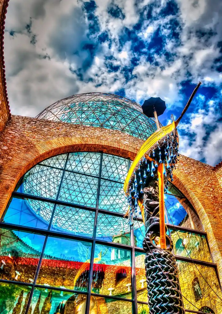 This image features the striking glass geodesic dome and brick archways of the Dalí Theatre-Museum in Figueres, Spain. A surreal sculpture of a boat dripping blue water-like shapes hangs dramatically in the foreground, reflecting Salvador Dalí's signature dreamlike aesthetic. The vivid sky and mirrored windows intensify the building's fantastical architecture.