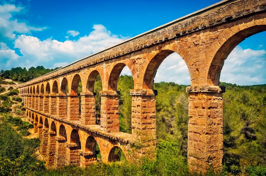 This image features an ancient Roman aqueduct made of warm-toned stone, stretching across a lush green valley under a bright blue sky. The structure is composed of two levels of evenly spaced arches, showcasing impressive engineering and symmetry. Its well-preserved condition highlights its historical significance and enduring beauty.