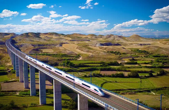 A sleek white high-speed train blurs across a long elevated viaduct, cutting through a vast rural landscape of rolling hills and patchwork farmland under a bright blue sky. The modern bridge and train contrast with the natural, open terrain, emphasizing efficient travel through remote areas.