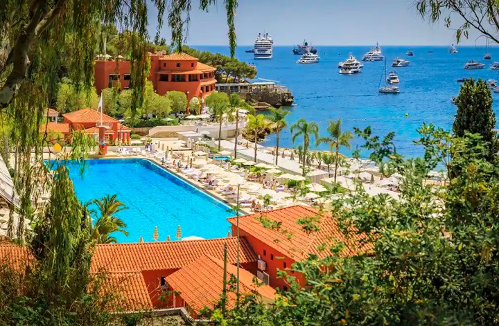 Thanks! Here's the SEO-optimized alt text based on your keyword: A luxury 5-star hotel in Monaco with terracotta rooftops, a large oceanfront pool surrounded by palm trees and sun loungers, overlooking the Mediterranean Sea dotted with yachts. The resort is nestled among lush greenery with elegant buildings and panoramic views of the coast.