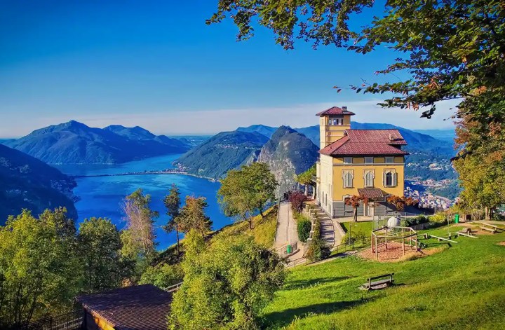 A scenic hilltop villa overlooks the deep blue waters of Lake Lugano, surrounded by lush green trees and dramatic mountain ranges under a bright blue sky. A walking path, benches, and a small playground sit in the foreground, enhancing the peaceful and inviting atmosphere.