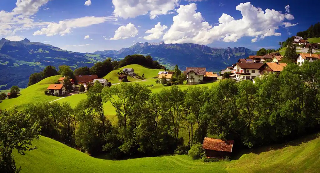 A panoramic view of a quaint alpine village nestled in lush green hills, with scattered traditional houses and red-roofed barns. Towering mountains loom in the background under a sky dotted with dramatic clouds, creating a serene and scenic landscape.