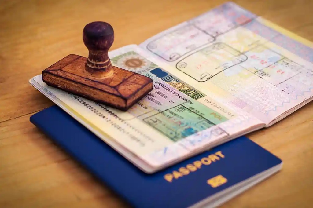 A wooden stamp rests on an open passport displaying a Schengen visa and various entry stamps. The passport with a dark blue cover lies on a wooden surface, symbolizing international travel and border control.