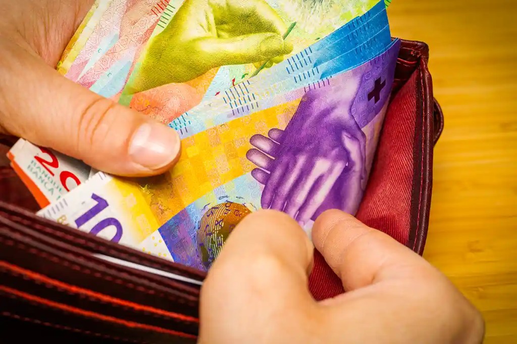 Close-up of hands holding a wallet filled with colorful Swiss franc banknotes in denominations of 10, 20, 50, and 100. The vibrant notes feature artistic hand illustrations and security elements, highlighting Swiss currencyโs distinctive design.