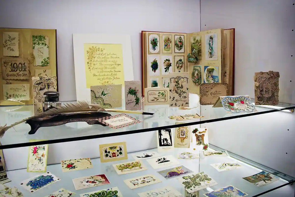 A glass display case showcases a collection of vintage greeting cards and ornate handwritten notes, some decorated with floral patterns and illustrations. A feather quill and ink bottle rest in front of the cards, evoking a nostalgic, old-world writing and gifting tradition.