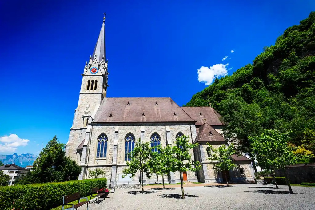 A stone Gothic-style church with a tall spire and stained glass windows stands under a vivid blue sky, nestled beside a lush green hillside. The courtyard is lined with young trees and benches, creating a peaceful and picturesque setting.