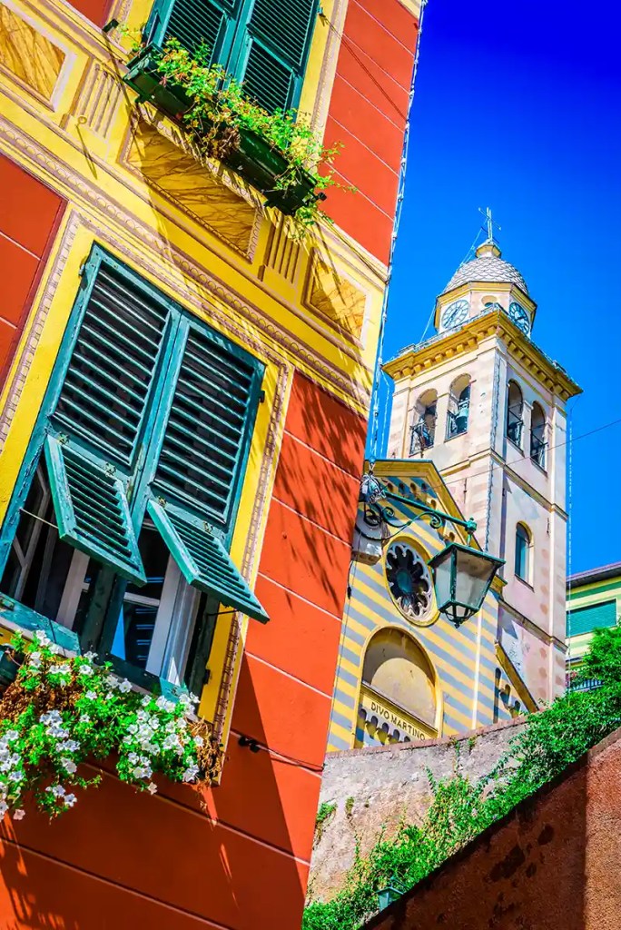 A brightly painted Italian building with green shutters and blooming flower boxes in the foreground, with the striped facade and clock tower of the Church of San Martino in Portofino rising behind it. The vivid colors pop under a clear, deep blue sky.