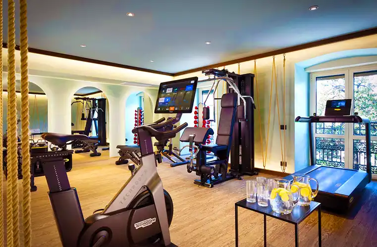 Modern hotel gym with Technogym equipment including stationary bikes, a treadmill, and a multi-station weight machine, set in a bright room with large windows and mirrors. A refreshment table with lemon water and glasses adds a welcoming touch.