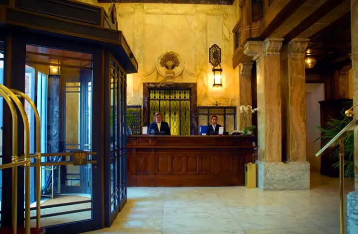 Two hotel receptionists stand behind a grand, vintage-style wooden check-in desk in a luxurious lobby with marble floors and ornate architectural details. A revolving door and luggage cart are visible in the foreground, highlighting the classic charm and upscale service of the hotel.