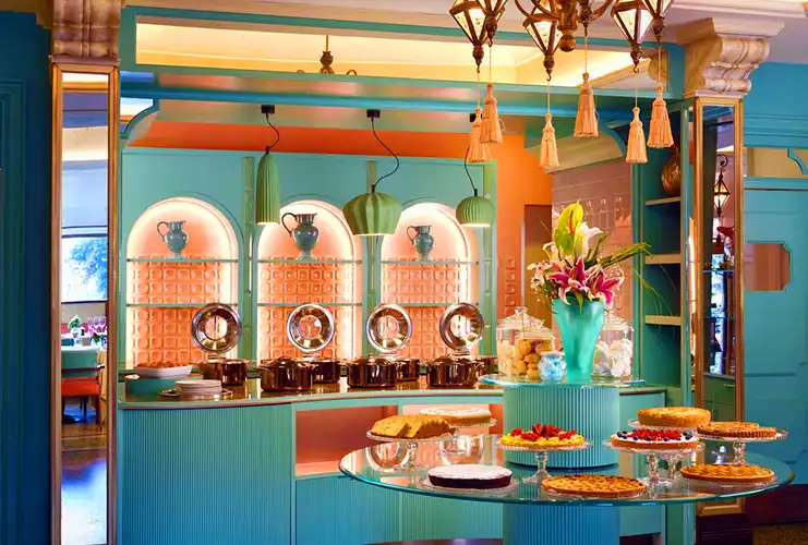 Colorful hotel breakfast buffet with a retro-inspired teal and coral design, featuring a variety of cakes, tarts, and pastries on a central glass table. Copper chafing dishes line the back counter beneath pendant lights, and a vase of fresh flowers adds a bright, elegant touch.
