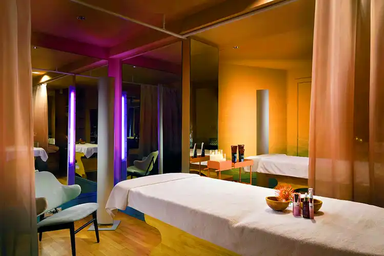 A softly lit spa treatment room with a massage table covered in a white sheet, surrounded by mirrors, blush-colored curtains, and ambient purple and amber lighting. Small bowls and skincare products are neatly arranged at the foot of the table, creating a serene, relaxing atmosphere.
