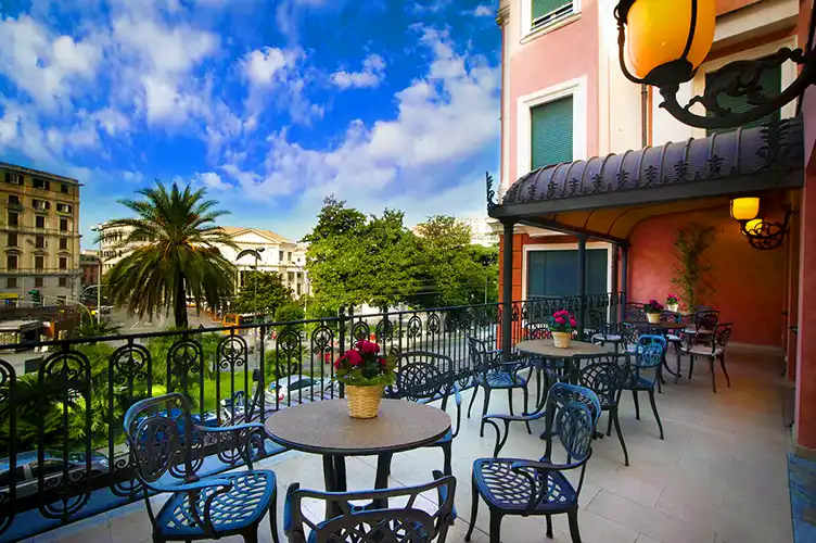 Charming hotel terrace with wrought iron tables and chairs, each decorated with a potted plant, overlooking a lush city square with palm trees and historic buildings under a bright blue sky. The coral-toned building and ornate railings add to the elegant Mediterranean vibe.