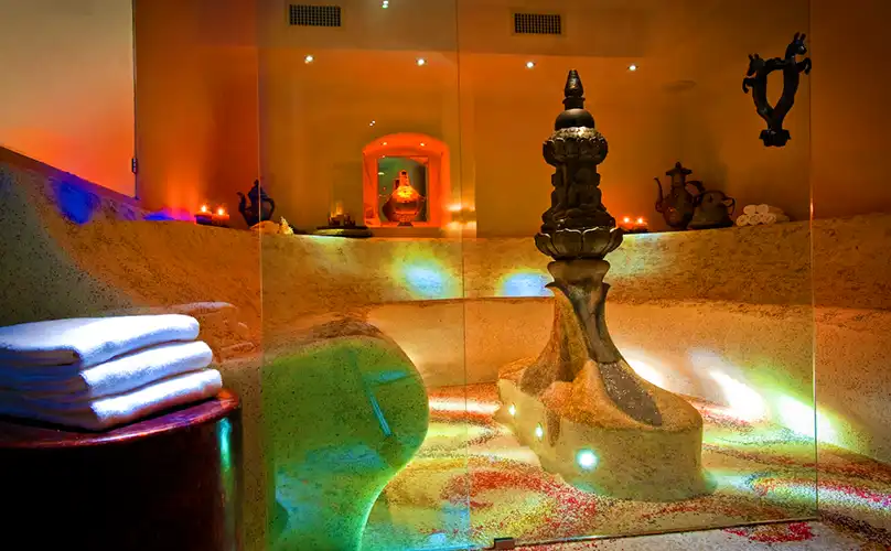 Luxurious spa steam room with curved stone seating, a central sculptural fountain, and colorful ambient lighting reflecting off the misty surfaces. Rolled white towels and ornate decorative vases enhance the warm, tranquil setting.