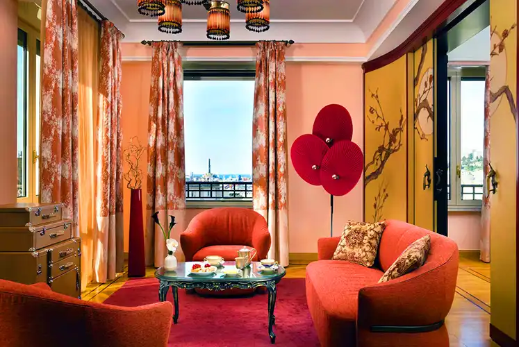 Vibrant hotel suite with red and coral tones, featuring plush seating around a coffee table set for tea and dessert. The space is decorated with floral curtains, painted folding screens, and unique red fan-shaped floor lamps, with city views visible through the windows.
