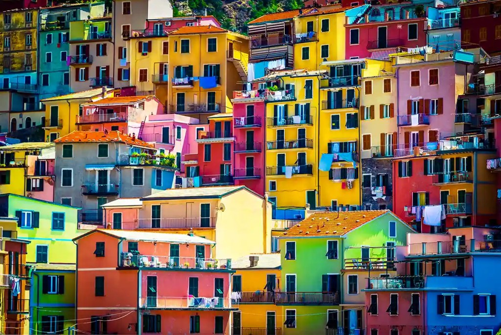 A vibrant hillside of colorful houses tightly stacked in the Italian village of Manarola, Cinque Terre. The buildings are painted in bold shades of yellow, red, pink, green, and orange, with balconies, laundry hanging out to dry, and dark green shutters.
