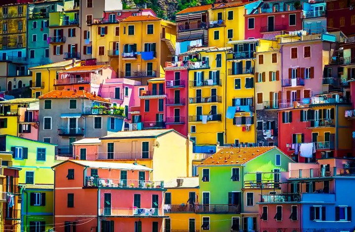 A vibrant hillside of colorful houses tightly stacked in the Italian village of Manarola, Cinque Terre. The buildings are painted in bold shades of yellow, red, pink, green, and orange, with balconies, laundry hanging out to dry, and dark green shutters.