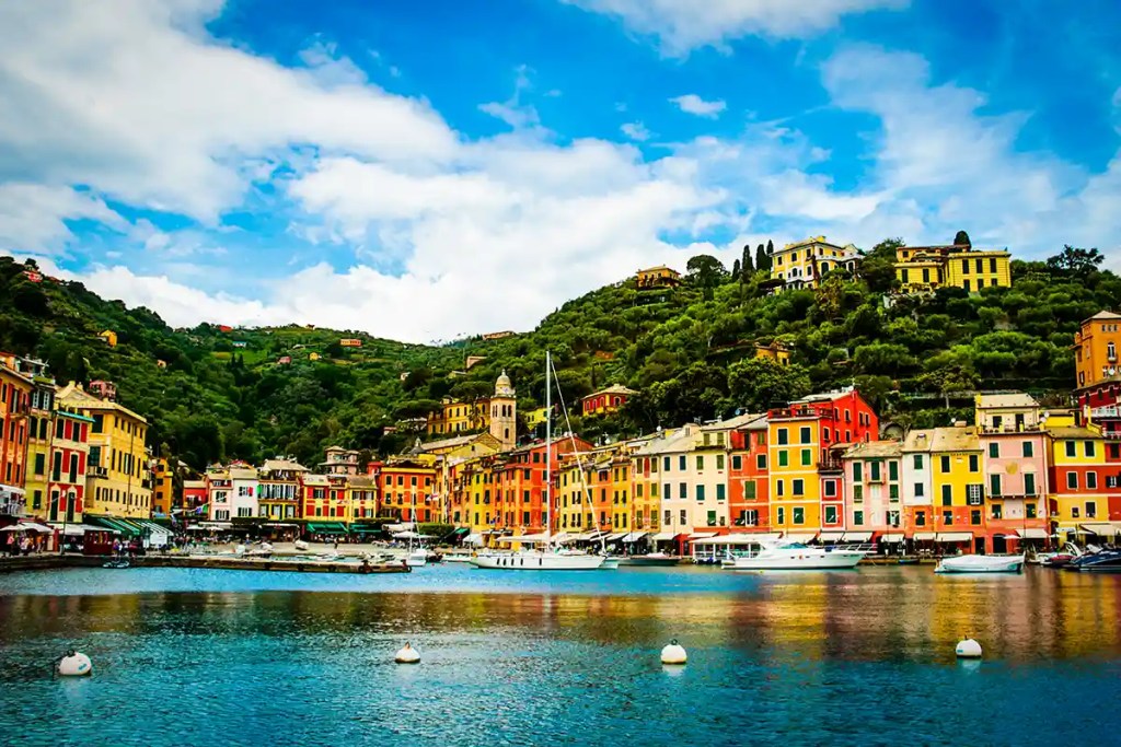 A scenic harbor in Portofino, Italy, with colorful buildings lining the waterfront and green hills rising in the background. Boats and yachts float on the calm blue water under a bright sky with scattered clouds.