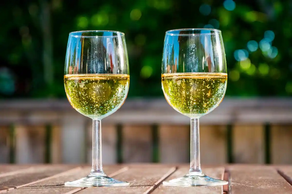 Two glasses of sparkling white wine sit on a wooden table outdoors with bubbles rising through the pale golden liquid. A softly blurred green background suggests a garden setting, creating a relaxed and celebratory atmosphere.