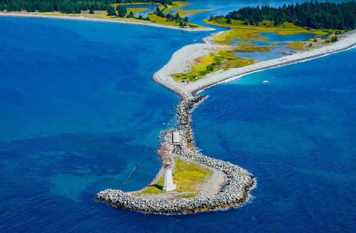 Alt text. Aerial view of a narrow rocky breakwater leading to a small white lighthouse surrounded by deep blue ocean and winding coastal inlets. The sweeping coastline and overhead perspective mirror the scenic views captured on a halifax helicopter tour.