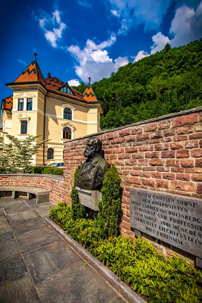 Bronze bust statue mounted against a curved brick wall beside a plaque that reads "JOSEF GABRIEL VON RHEINBERGER 1839 VADUZ 1901 MUNICH". Behind the memorial stands a pale yellow historic building with decorative towers and a forested mountainside under a bright blue sky with scattered clouds.