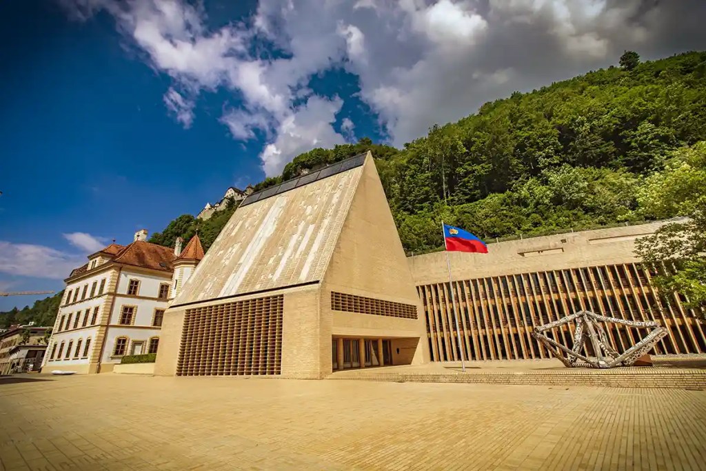 Modern angular stone building with a steep triangular roof beside a long columned structure and the Liechtenstein flag flying in front. A historic cream colored building and a forested hillside rise behind the plaza under a dramatic sky filled with large white clouds.