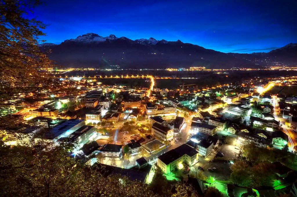 Wide view of a town at dusk with glowing streetlights and illuminated buildings spread across the valley floor. Snow capped mountains form a dark silhouette against a deep blue evening sky, while winding roads create bright lines of light through the city below.