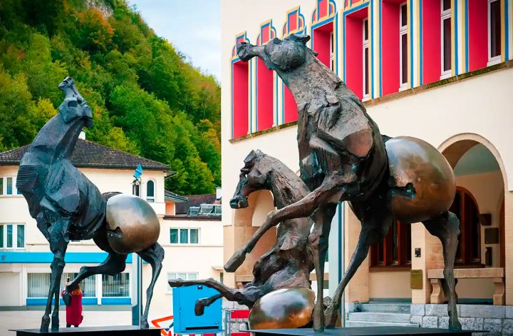 Two large bronze horse sculptures balance with metallic spheres near their hind legs in a public square in Liechtenstein. The dramatic statues stand in front of a colorful building with red window frames and a forested hillside behind them.