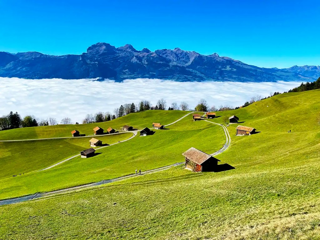 Rolling green alpine hills dotted with small wooden barns connected by winding farm roads. A thick layer of clouds fills the valley below while rugged mountains rise above the mist under a deep blue sky.
